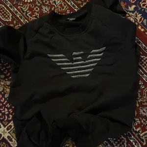 Svart sweatshirt från Armani - Svart sweatshirt från Armani med det ikoniska silverfärgade örnloggan på bröstet. Tröjan har rund hals och långa ärmar, perfekt för en clean och stilren look. Materialet känns mjukt och skönt mot huden.