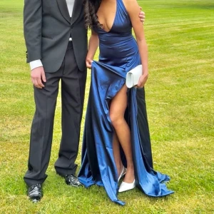 Marinblå långklänning med slits - Säljer en elegant marinblå långklänning i glansigt satinmaterial. Klänningen har djup v-ringning, är ärmlös och har en hög slits på ena sidan som ger en snygg och modern look. Perfekt för bal eller fest. Passformen är slim och tyget faller snyggt längs kroppen.