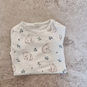 Vit långärmad body med björnar från Newbie - Supergullig långärmad vit body från Newbie i storlek 74, med ett sött mönster av bruna björnar och blå blad. Tröjan är gjord i mjuk bomull och har praktiska tryckknappar vid axeln för enkel påklädning. Perfekt för små äventyrare! Plagget är inköpt ny och sparsamt använd av ett bra. 