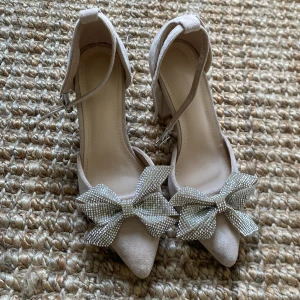 Beige klackskor med glittriga rosetter - Snygga beige klackskor med spetsig tå och glittriga rosetter framtill. Skorna har en bekväm blockklack och justerbara remmar runt ankeln. Perfekta för att lägga till lite glamour till din outfit.