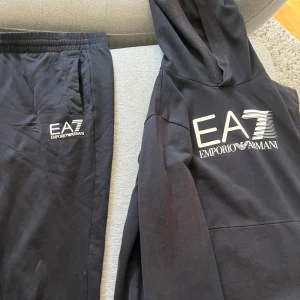 Svart EA7 Emporio Armani huvtröja - Svart huvtröja från EA7 Emporio Armani med stor vit logga på bröstet.  Byxorna har använts en del jämfört med hoodien