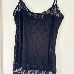 Svart spetsig linnetopp med rosett - Svart linnetopp med tunna axelband och helspets i ett snyggt blommigt mönster. Toppen har en liten rosett framtill och är genombruten med transparenta detaljer. Perfekt för dig som gillar en feminin och trendig look.