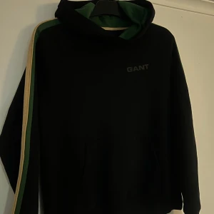 Svart hoodie från GANT! - Svart hoodie från Gant med grön/beiga ränder längst armarna. Tröjans huva har en grön insida och är i strl 170/176 Riktigt snygg! Mkt fint skick!
