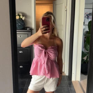 Rosa bandeau tubtopp med rosett - Superfin rosa bandeau tubtopp med stor rosett framtill och volangkant upptill. Toppen är axelbandslös och har en luftig, lätt utsvängd passform som ger en söt och trendig vibe. Perfekt för varma dagar och sommarhäng.