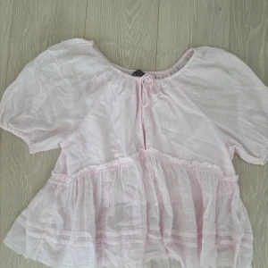 Ljusrosa blus från Amelie & Me - Supersöt ljusrosa blus med puffiga korta ärmar och volangdetaljer från Amelie & Me. Blusen har en rund halsringning med knyt framtill och är gjord i ett tunt, luftigt bomullstyg som ger en romantisk vibe. Perfekt för vår och sommar!