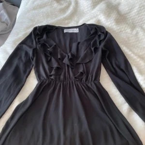 Svart volangklänning Ivyrevel - Supersnygg svart klänning från Ivyrevel med långa ärmar och v-ringad hals. Volangdetaljer framtill ger en romantisk vibe och midjan är lätt rynkad för en smickrande siluett. Perfekt för dig som gillar stilrena plagg med lite extra edge.