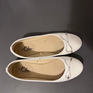 Vita ballerinaskor med rosett Clou - Snygga vita ballerinaskor från Clou i storlek 36 (MEN PASSAR 37!! Stor i storleken) Skorna har rund tå, platt sula och en söt rosett framtill med silvriga detaljer. Perfekt för dig som gillar enkel och stilren design. Materialet är syntet och insidan är ljusbeige. Använt fåtal gånger, i mycket bra skick!