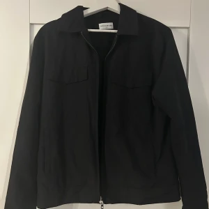 Svart overshirt från These Glory Days - Stilren svart overshirt från These Glory Days med klassisk krage, två bröstfickor med lock och dragkedja framtill. Jackan har lång ärm och en clean look som passar till allt. Perfekt för lager-på-lager och enkel att matcha.