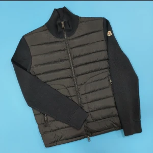 MONCLER CARDIGAN  - Snygg svart pufferjacka från Moncler med quiltad framsida och stickade ärmar. Jackan har hög krage, dragkedja framtill och två fickor med dragkedja. Klassisk Moncler-logga på ärmen och ribbade muddar för extra stilpoäng. Pris kan diskuteras 