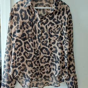 Leopardmönstrad skjorta från SHEIN - Snygg leopardmönstrad skjorta från SHEIN i tunn och luftig polyester. Skjortan har klassisk krage, två bröstfickor och knappar framtill. Färgerna går i beige och svart, perfekt för dig som vill sticka ut med ett djuriskt print.