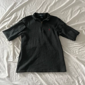 Svart pikétröja från Polo Ralph Lauren - Svart pikétröja från Polo Ralph Lauren med kort ärm och half zip-dragkedja framtill. Tröjan har en klassisk krage och röd broderad logga på bröstet. Tillverkad i 100% bomull för skön och mjuk känsla. Perfekt för dig som gillar stilrena och tidlösa plagg. Funkar bättre storlek M.