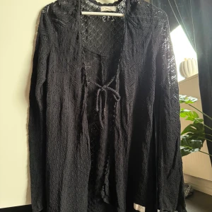 Svart spetsväst från Odd Molly - Svart lång väst från Odd Molly med snyggt spetsmönster och knytning framtill. Västen har ett genomarbetat geometriskt mönster och är tillverkad i ett tunt, stickat material som ger en cool och bohemisk vibe. Perfekt att slänga över en t-shirt eller klänning.