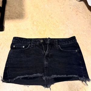 Svart jeanskjol med fransig kant - Cool svart jeanskjol med rå, fransig kant och klassiska fickor både fram och bak. Kjolen har bälteshällor och dragkedja framtill. Perfekt för dig som gillar en edgy och avslappnad stil.