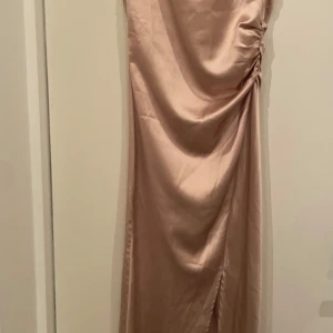 Champagnefärgad klänning Neo Noir - Säljer en elegant klänning från Neo Noir i champagnefärgad satin. Klänningen är ärmlös med draperad detalj och snörning på sidan, samt har en diskret slits. Perfekt för dig som gillar stilrena och lyxiga vibes.