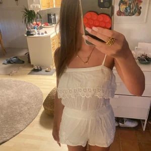 Vit onesuit med spetsdetaljer - Säljer en superfin vit onepize med tunna axelband och snygga spetsdetaljer både upptill och nedtill. Plagget är ärmlös och har en luftig, lätt struktur som passar perfekt för varma dagar. Spetsen ger en romantisk och bohemisk vibe. Axelbanden knyts med rosett på axeln.