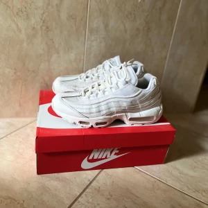 Nike Air Max 95 Essential vit - Nike Air Max 95 Essential sneakers i fräsch vit och ljusgrå färg. Ikonisk design med synliga Air Max-enheter i sulan, mesh och syntetmaterial på ovandelen samt snygga vågiga paneler. Skorna har snörning och rund tå, perfekt för dig som gillar streetwear och sportig stil.