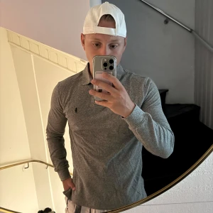 Grå pikétröja från Polo Ralph Lauren - Snygg grå långärmad pikétröja från Polo Ralph Lauren i custom fit. Tröjan har klassisk krage, knappar vid halsen och den ikoniska broderade loggan på bröstet. Perfekt för dig som gillar stilrena och tidlösa plagg.