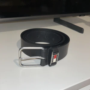 Svart skärp från Tommy Jeans i skinn - Snyggt svart bälte från Tommy Jeans i äkta skinn med klassiskt silverfärgat spänne. På hällan sitter den ikoniska Tommy Jeans-loggan i rött, vitt och blått. Perfekt accessoar för att lyfta din outfit med en clean och stilren look.