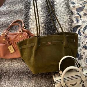 Olivgrön mocka axelväska - Säljer en olivgrön axelväska i mocka med stilrena linjer och långa handtag. Väskan har en enkel design med dekorativt band och metallspänne på framsidan. Perfekt storlek för att få plats med allt du behöver och passar till många olika outfits. Lite sliten och smutsig men ser till att rengöra innan jag fraktar!