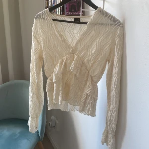 Krämvit volangblus från Gina Tricot - Supersöt krämvit blus från Gina Tricot med lång ärm, v-ringning och volangdetalj framtill. Tunt, lätt transparent tyg med struktur och vågigt mönster. Perfekt att styla med jeans eller kjol för en trendig look💕