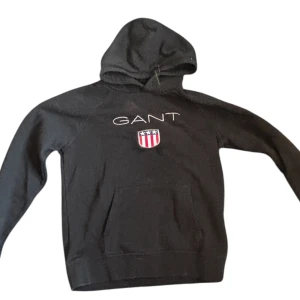 Svart hoodie från GANT med logga - Svart hoodie från GANT med broderad logga och emblem på bröstet. Tröjan har huva, magficka och långa ärmar. Tillverkad i mjuk bomullsmix som är skön att ha på sig. Perfekt för dig som gillar klassisk streetstyle och snygga detaljer.