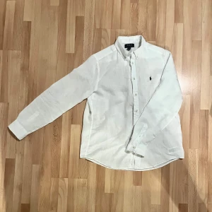 Vit linneskjorta från Polo Ralph Lauren - Snygg vit skjorta från Polo Ralph Lauren i linne med klassisk krage och knappar framtill. Långärmad modell med mörkblå broderad logga på bröstet. Perfekt passform som sitter normal och känns lätt och fräsch, grym till jeans eller shorts. Använd en gång. Storleken motsvarar S/M