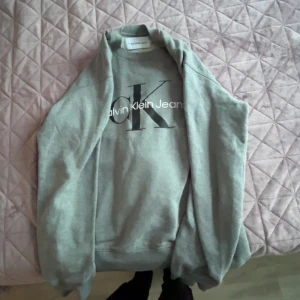 Grå Calvin Klein Jeans sweatshirt - Säljer en grå sweatshirt från Calvin Klein Jeans med stort CK-tryck på bröstet. Tröjan har rund hals, långa ärmar och ribbade muddar. Mjuk insida och klassisk passform, perfekt för chill dagar. Storlek M.