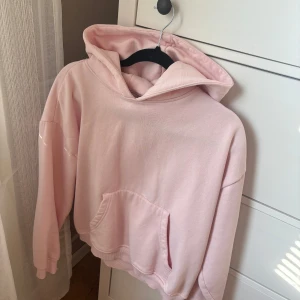 Ljusrosa hoodie med huva - Säljer en ljusrosa hoodie med stor huva och magficka. Tröjan har en loose passform och långa ärmar, perfekt för chill dagar. Materialet är mjuk bomull som känns skönt mot huden. Enkel och stilren design utan tryck eller mönster.