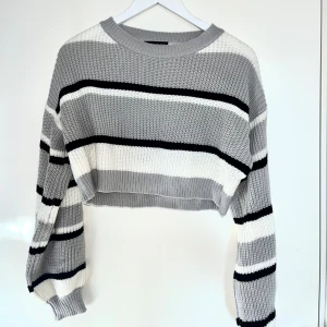 Randig croppad stickad tröja Shein - Säljer en croppad stickad tröja från Shein med breda ränder i grått, vitt och svart. Tröjan har långärmade ärmar, rund hals och ribbade muddar. Perfekt för dig som gillar chill och trendig stil med lite loose passform.