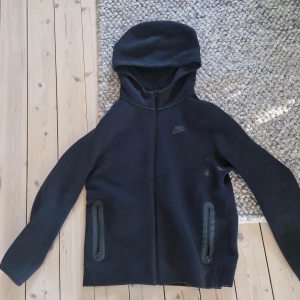 Svart Nike tech fleece gen 2 XL kids - Svart hoodiejacka från Nike i storlek XL, med hel dragkedja och två fickor med dragkedja. Jackan har en stilren look med diskret Nike-logga på bröstet och är tillverkad i mjuk polyester. Perfekt för chill dagar eller när du vill ha en sportig vibe. Är typ använda 5 gånger. Finns byxor som hör till på mitt konto.