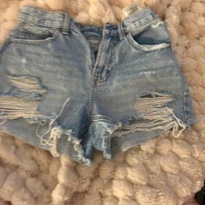 Blå slitna jeansshorts med fransar - Säljer ett par ljusblå jeansshorts fransiga kanter. Shortsen har slitna detaljer och hål framtill, knappt använda från Vermoda