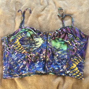 Färgglad korsettopp med dragkedja - Säljer en unik korsettopp med färgstarkt abstrakt mönster i blått, lila, gult och grönt. Toppen har smala axelband och en dragkedja framtill. Perfekt för dig som vill sticka ut med en cool och edgy look.