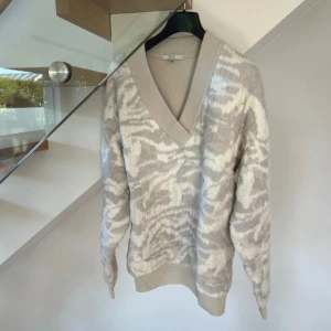 Beige mönstrad stickad tröja Dagmar - Säljer en beige och vit stickad tröja från Dagmar med snyggt abstrakt mönster. Tröjan har bred ribbad v-ringad krage, långa ärmar och ribbade muddar. 