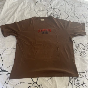 Brun Champion t-shirt med tryck - Snygg brun t-shirt från Champion i 100% bomull. Klassisk passform med korta ärmar och rund hals. Framsidan har ett rött och svart Champion-tryck med japanska tecken. Perfekt för dig som gillar streetwear och vill ha en chill look. Inga defekter. Priset kan diskuteras. 