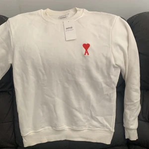 Vit sweatshirt från Ami med rött hjärta - Vit sweatshirt från Ami med klassisk rund hals och lång ärm. På bröstet finns ett broderat rött hjärta med bokstaven A. Tröjan är gjord i mjuk bomull och har ribbade muddar vid ärmslut och nederkant. Perfekt för dig som gillar stilrena och trendiga plagg. Lite smutsig men går bort i tvätten 