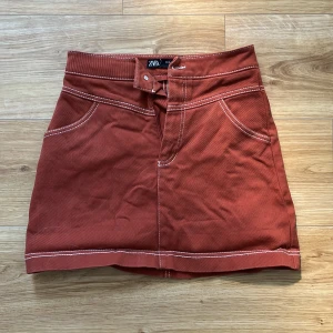 Kjol Zara - Kort mörkorange kjol från Zara med vita sömmar. Dragkedja och knapp i gylfen. Två fickor på framsidan, inga på baksidan. Storlek: XS. Material: 99% bomull, 1% elastan. Sparsamt använd, i bra skick!
