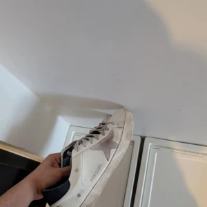 Golden Goose Superstar sneakers - Säljer ett par Golden Goose Superstar sneakers i vitt med grå stjärndetalj på sidan och mörkblå häl. Skorna har grå snörning, rund tå och platt sula. Materialet är skinn och mocka, med klassisk Golden Goose-design och coola detaljer. Det är äkta men vill bara till lägga att det är lite nött på sulan under hällen som det såklart blir på sånna här skor🙄men övrigt så är det en utmärkt och snygg sko till ett relativt bra pris 😌priset är ej hugget i sten 🪨 🗡️ 