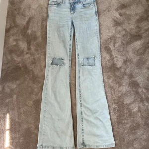 Bootcut jeans med slitna knän från Nelly,  - Snygga ljusblå bootcut jeans från Nelly med slitna hål på båda knäna och coola röda broderade detaljer på bakfickorna. Jeansen har klassisk femficksdesign, knapp och dragkedja framtill samt kontrastsömmar. Perfekt för dig som gillar retrovibe och statement-jeans.