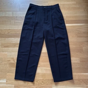 Svarta kostymbyxor från Bershka - Snygga svarta kostymbyxor från Bershka med klassisk rak passform och pressveck framtill. Byxorna har midja med knapp och dragkedja, samt bälteshällor och fickor både fram och bak. Perfekta för en clean och stilren look.