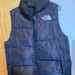 Svart dunväst från The North Face S - Svart dunväst från The North Face med hög krage och dragkedja framtill. Västen har två fickor med dragkedja och broderad logga på både bröstet och ryggen. Perfekt för lager-på-lager och riktigt clean look.