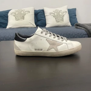Golden Goose Superstar sneakers med grå stjärna - Golden Goose Superstar sneakers i vitt skinn med grå stjärndetalj på sidan och svarta detaljer vid hälen. Skon har grå snörning och en klassisk platt sula. Ikonisk och stilren modell från Golden Goose, tillverkad i Italien.