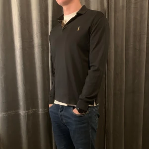 Svart pikétröja från Ralph Lauren - Snygg svart pikétröja från Ralph Lauren med klassisk krage och knappdetaljer. Tröjan har långa ärmar och en liten broderad logotyp på bröstet. Perfekt för en stilren look.