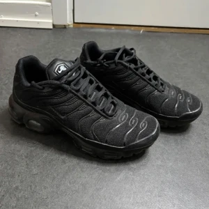 Svarta Nike Air TN sneakers - Säljer ett par helsvarta Nike Air TN sneakers med klassisk Tn-detalj på hälen. Skorna har mesh- och syntetöverdel, rund tå och tydliga vågformade linjer på ovandelen. Perfekta för dig som gillar streetwear och sportig stil.