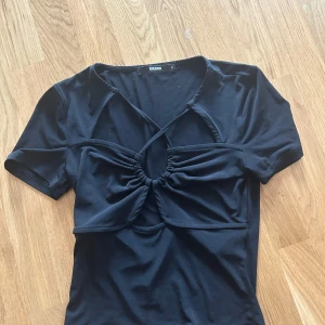 Svart croppad topp med knyt från BikBok - Säljer en svart croppad topp från BikBok med korta ärmar och snygga knytdetaljer framtill. Toppen har en unik design med rynk och öppen detalj vid bysten. Perfekt för att lyfta din outfit!
