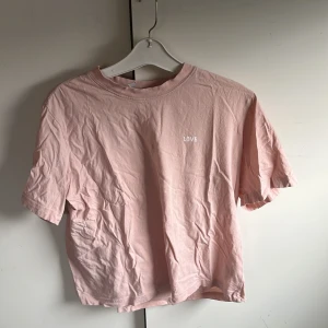 Ljusrosa t-shirt med trycket 'LOVE' från H&M  - Ljusrosa t-shirt från H&M med litet vitt tryck 'LOVE' på bröstet. Klassisk rund halsringning och korta ärmar. Tillverkad i mjuk bomull för en bekväm känsla.