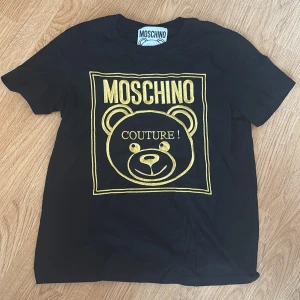 Svart t-shirt från Moschino - Snygg svart t-shirt från Moschino med ett stort guldtryck av en björn och texten 'Couture!' på framsidan. Perfekt för dig som vill ha en stilren men ändå iögonfallande look. T-shirten har en normal passform och korta ärmar. Är M men sitter som S