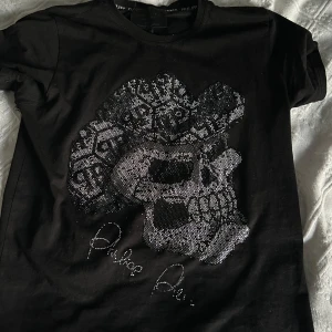 Svart t-shirt med dödskallemotiv från Philipp Plein - Snygg svart t-shirt från Philipp Plein med ett coolt dödskallemotiv i glittrande detaljer. Perfekt för att ge din outfit en edgy touch. T-shirten har korta ärmar och är gjord i ett bekvämt material.