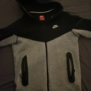Grå och svart hoodie från Nike - Säljer en snygg grå och svart hoodie från Nike med dragkedja och huva. Jackan har långa ärmar och praktiska fickor med dragkedja. Perfekt för en sportig look eller en avslappnad dag. Den är äkta från stadiuem.
