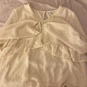 Vit klänning från Lindex - Säljer en söt vit klänning från Lindex med volangdetaljer och diskret prickigt mönster. Klänningen har långa ärmar och en liten knapp i nacken. Perfekt för speciella tillfällen eller en fin sommardag. 🌼