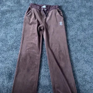Bruna velourbyxor från Juicy Couture - Säljer ett par bruna velourbyxor från Juicy Couture med strassdetaljer och Juicy-text baktill. Byxorna har dragsko i midjan och raka ben. Perfekta för en avslappnad och trendig look.